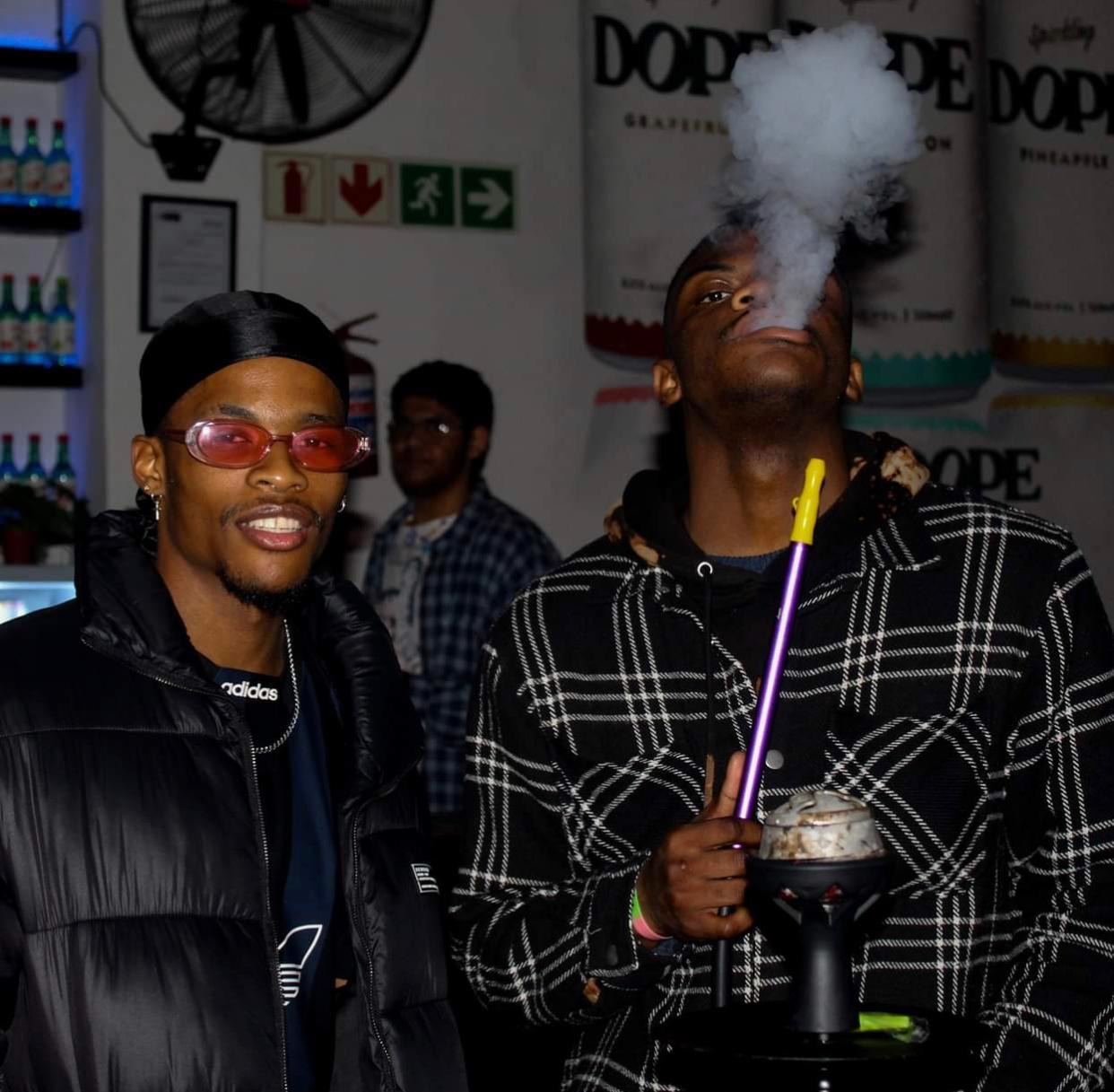 Two guests under warm venue light, one exhaling shisha smoke